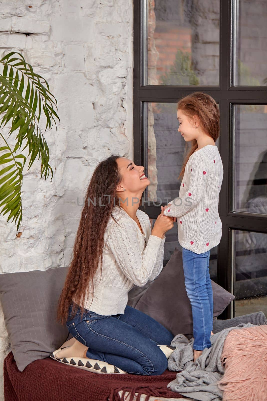 Mother and daughter sitting on sill near window in room. They show emotions and have fun