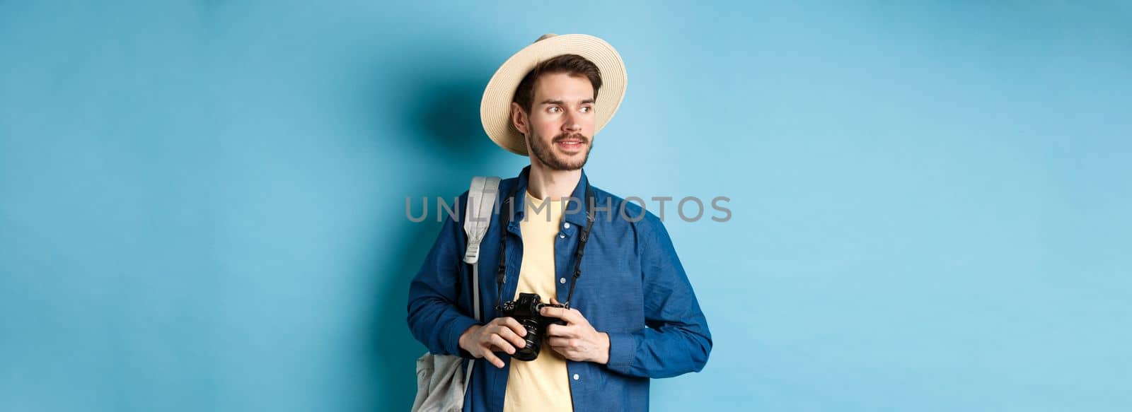 Handsome positive guy in summer hat, holding photo camera and looking aside, tourist taking pictures on vacation, standing with backpack on blue background.