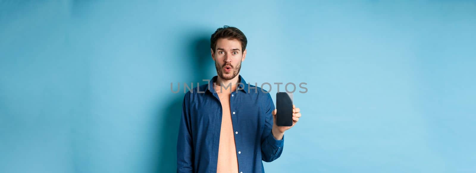 Impressed caucasian male model showing smartphone screen and look amazed, talking about online offer, standing on blue background.