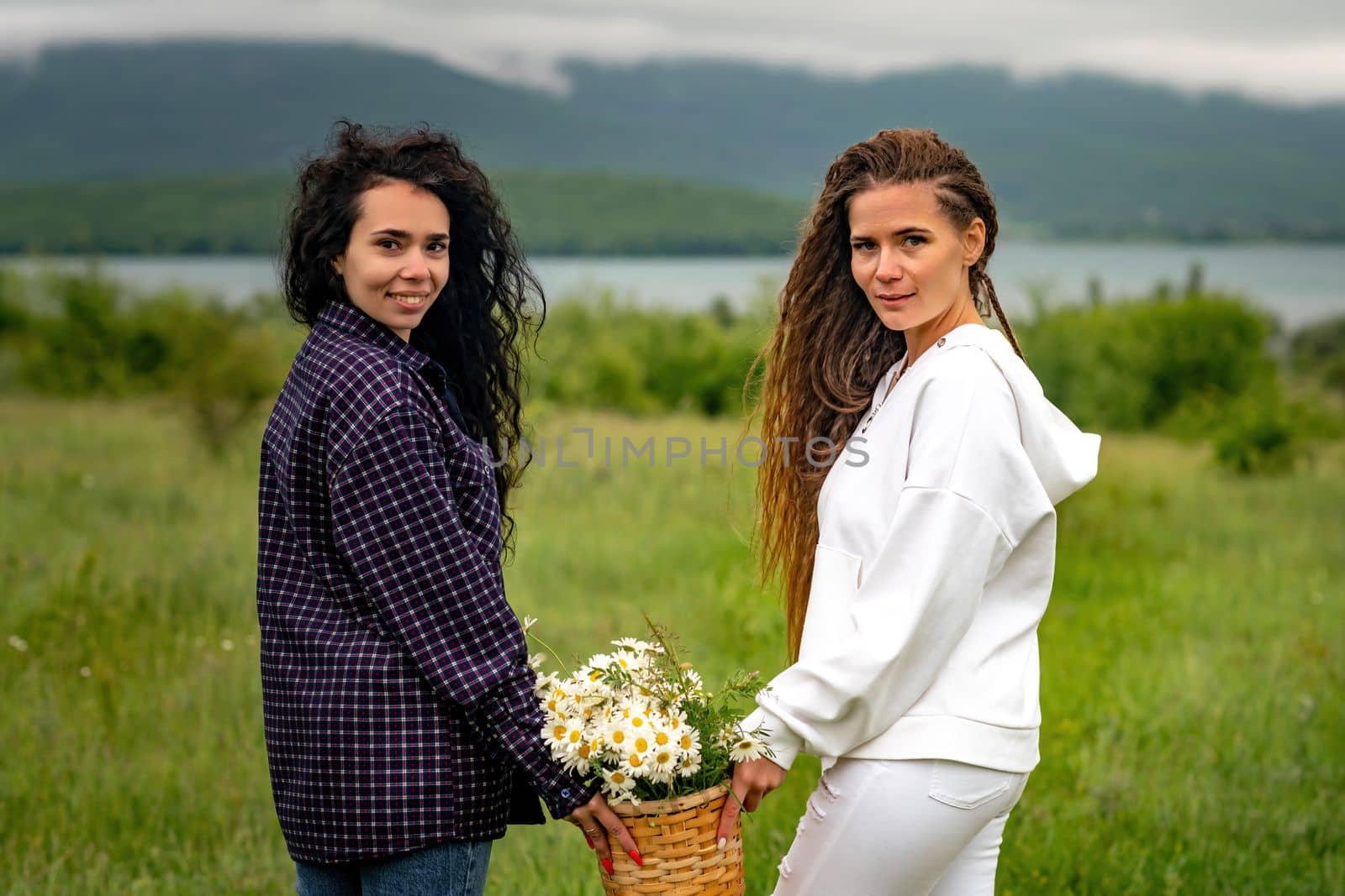 Two women enjoy nature in a field of daisies. Girlfriends hugging hold a bouquet of daisies and look at the camera. by Matiunina