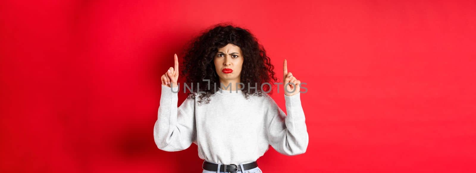 Grumpy young woman with curly hair frowning and grimacing unsatisfied, pointing fingers up at something bad, complaining on company, red background.