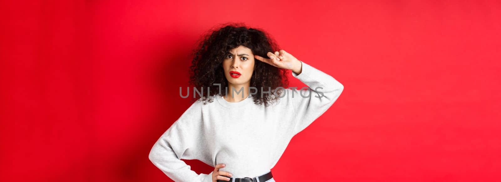 Are you stupid. Annoyed and confused girl with curly hair, pointing at head and scolding person being crazy or strange, standing on red background.