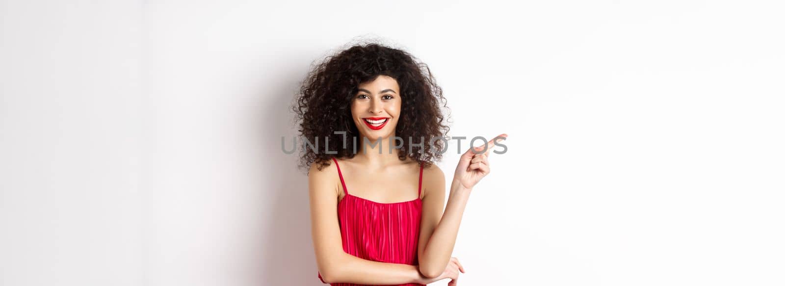 Beautiful happy woman with curly hairstyle, wearing festive dress, pointing finger left at logo and smiling, white background.