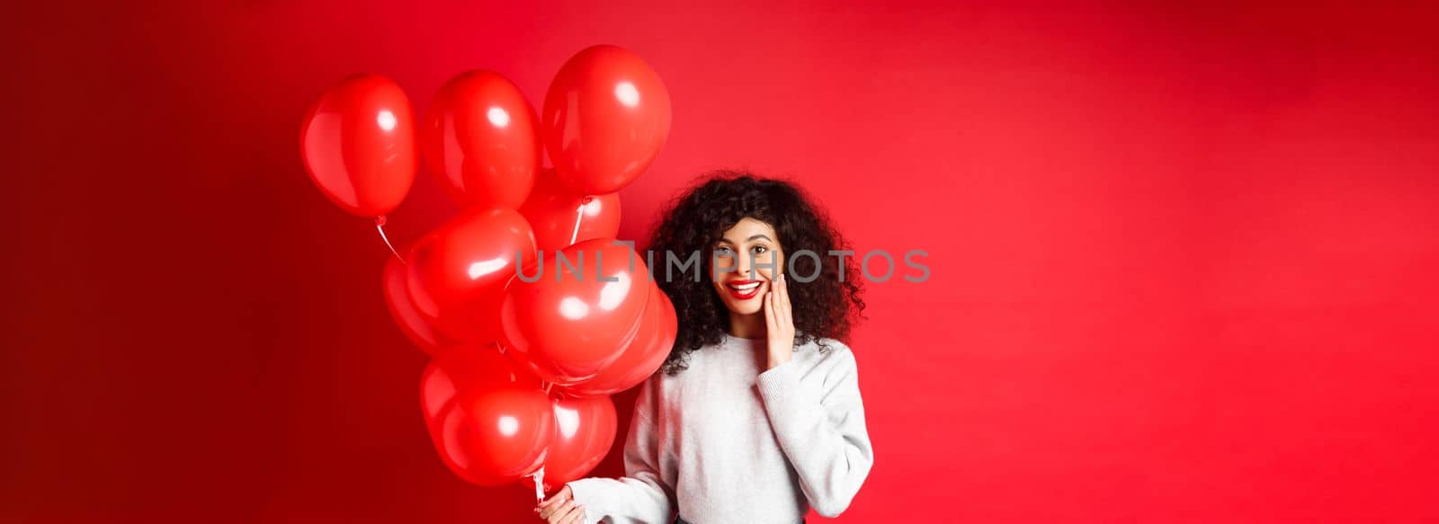 Holidays and celebration. Surprised birthday girl holding helium party balloons and looking touched at camera, being congratulated, red background.