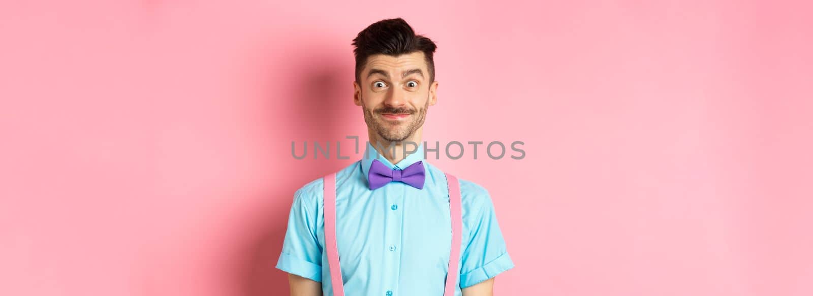 Smiling caucasian man looking excited, standing in classy bow-tie and shirt for romantic date, pink background.