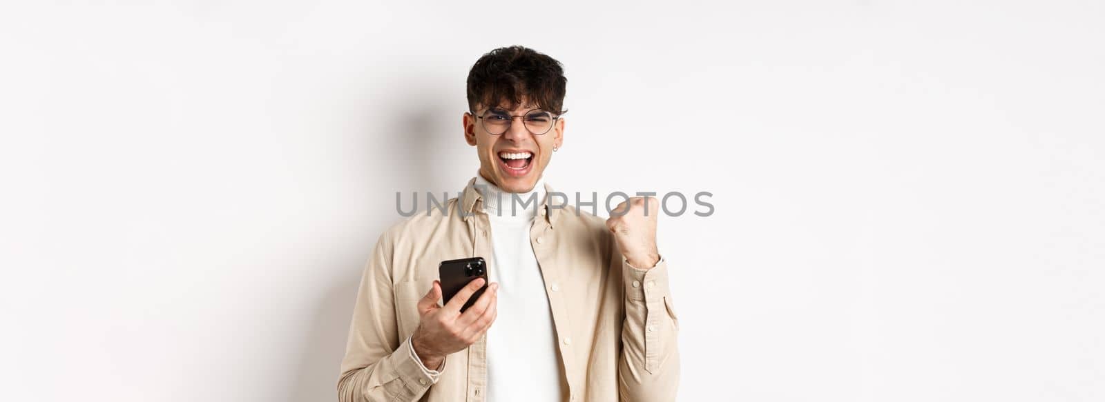 Technology and online shopping concept. Motivated and happy guy scream yes and show fist pump after winning on smartphone, achieve goal in app, white background by Benzoix