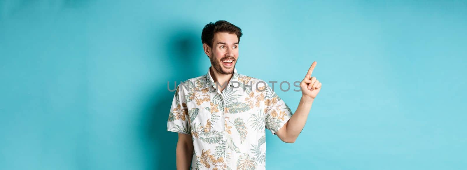Excited smiling tourist pointing and looking aside at empty space, checking out promo offer, standing in hawaiian shirt on blue background.