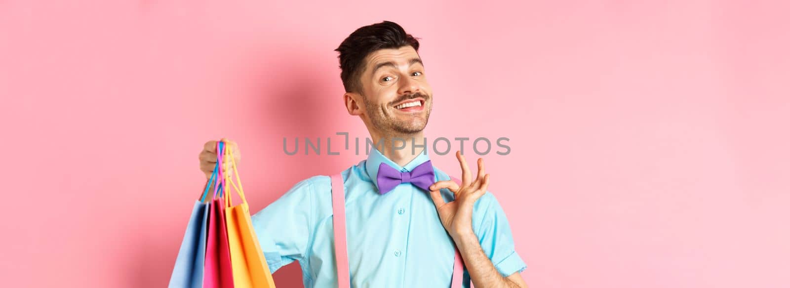 Confident smiling guy adjusting his bow-tie and showing gifts in shopping bags, looking happy, standing over pink background.