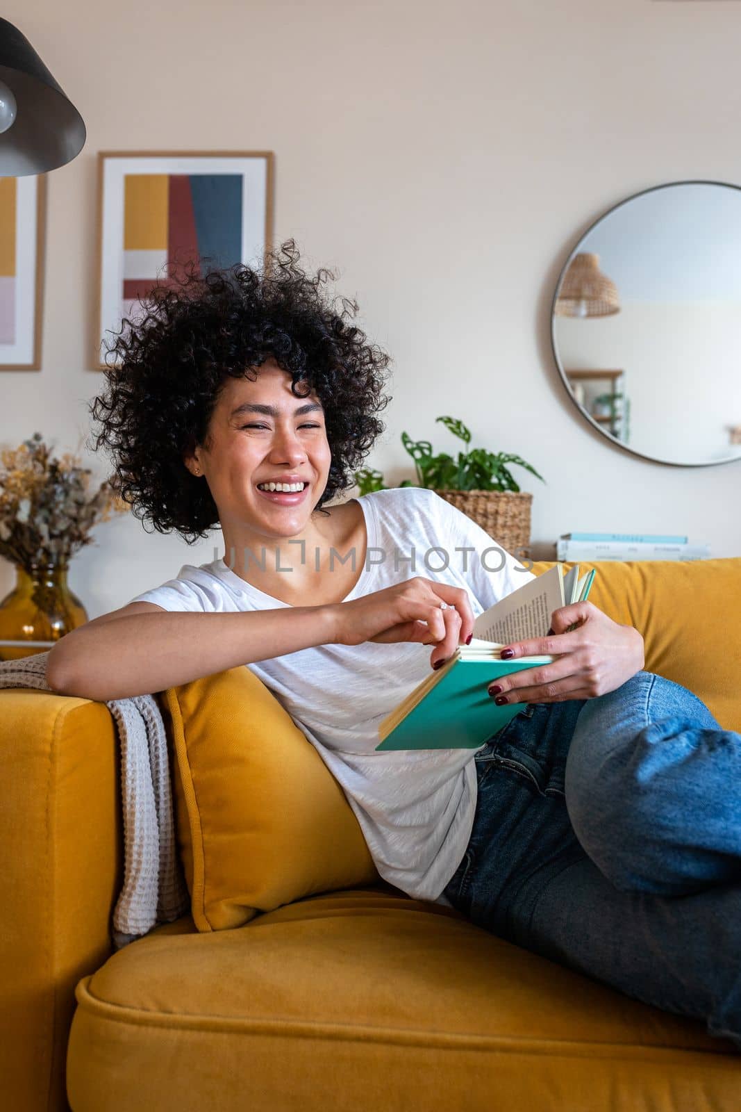 Portrait of happy, smiling African American woman relaxing at home reading a book sitting on the couch looking at camera. Vertical image. Lifestyle concept.