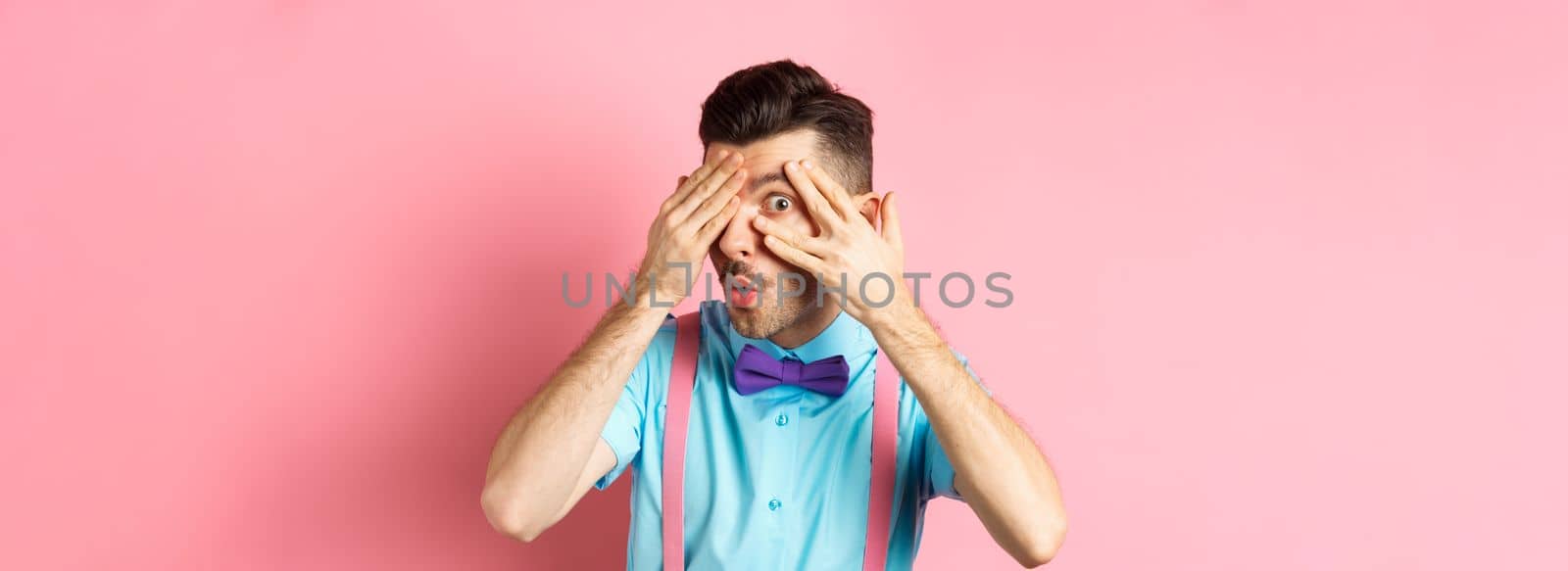 Intrigued funny guy in bow-tie, peeking through fingers at something interesting, checking out something cool, standing on pink background.