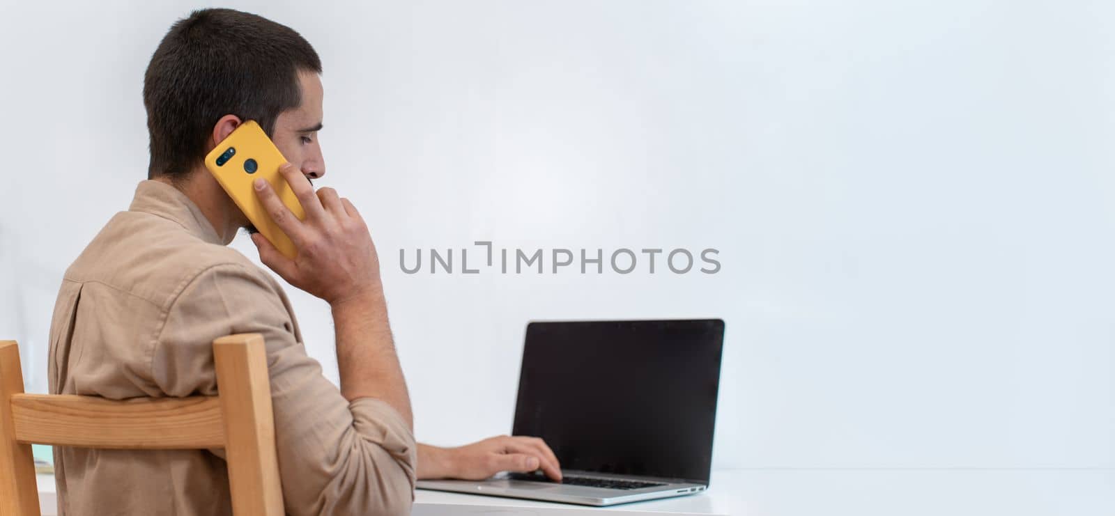 Young freelancer sitting at his work desk talking with smart phone call in front of his laptop. High quality photo with copy space for text banner.
