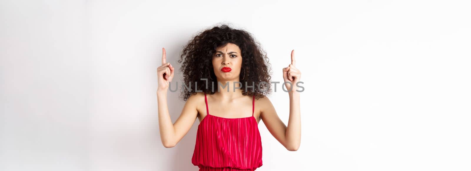 Disappointed frowning woman with makeup, wearing red dress on valentines, pointing fingers up and complaining, standing on white background by Benzoix