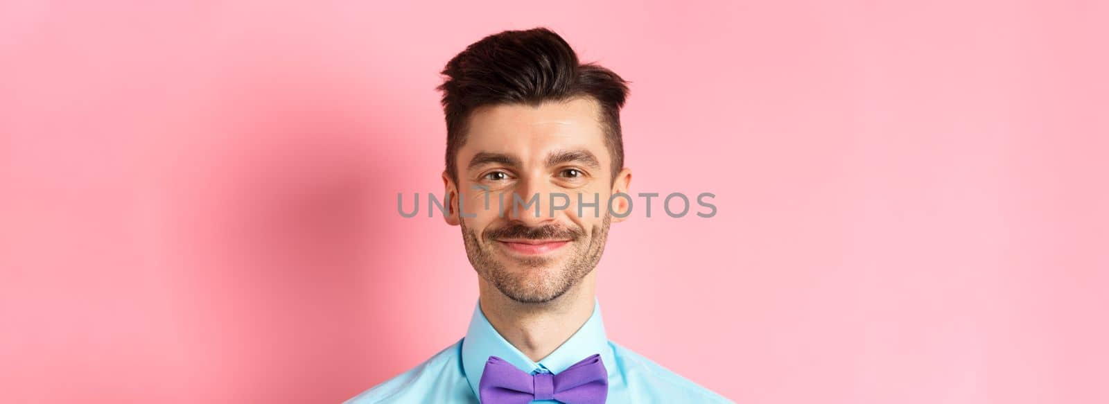 Close-up of smiling hansome man with moustache, wearing bow-tie and shirt, standing cheerful on pink background.