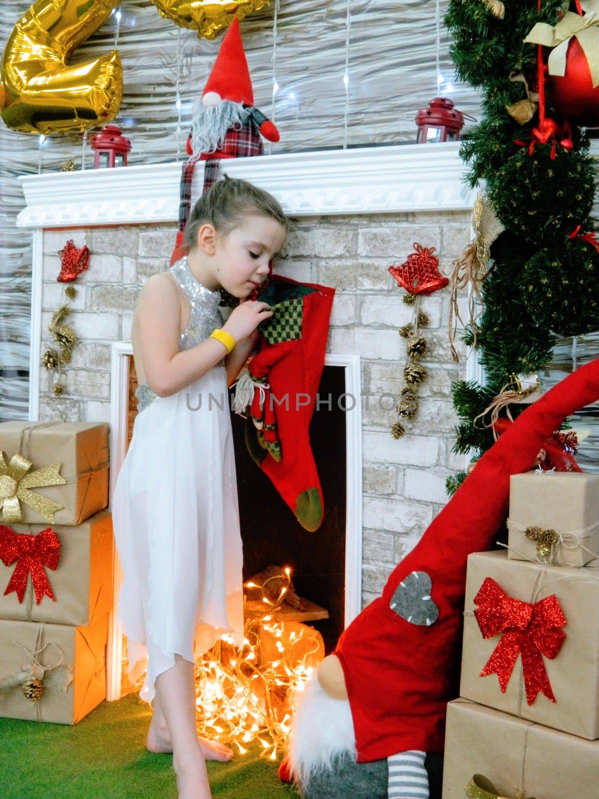 A little girl in holds a box of gifts and rejoices. The child sits near a Christmas tree and unpacks gifts by milastokerpro