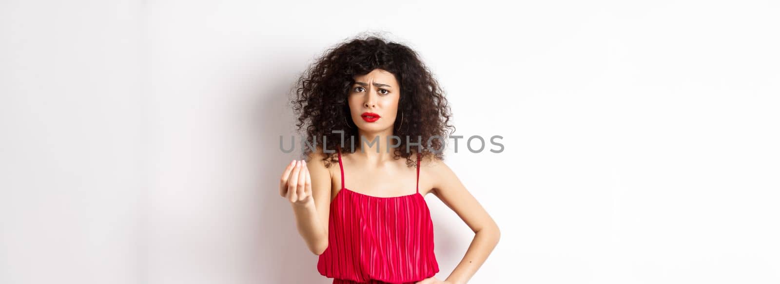 Confused woman with curly hair, wearing red dress, frowning and shaking hand, cant understand something, looking hesitant, standing on white background.