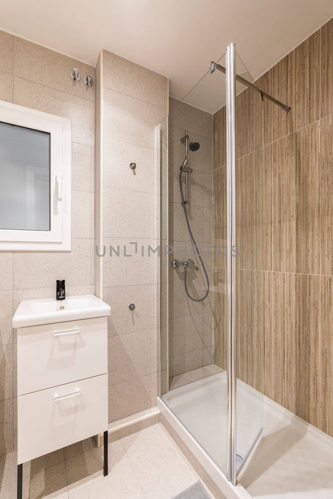 Corner of bathroom with small shower area enclosed by transparent glass wall. Washbasin on table with drawers for bathroom accessories. Window in wall with frosted glass and white frame