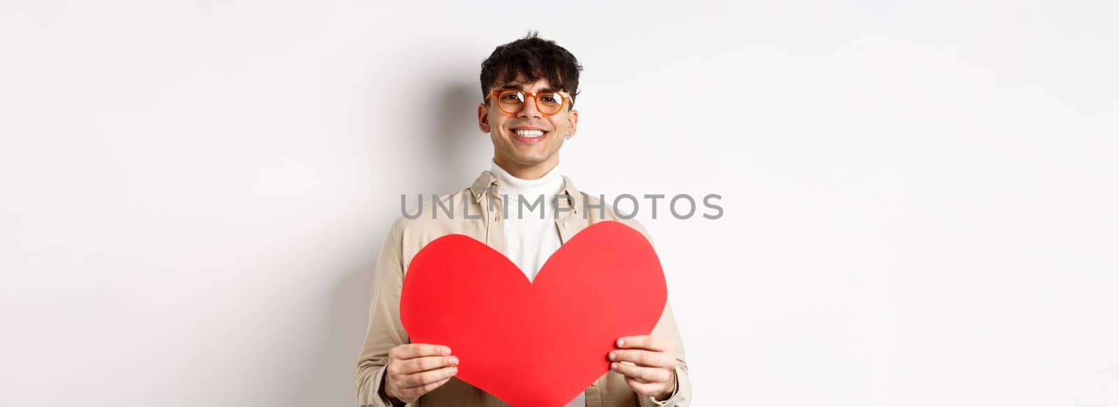 Attractive modern man smiling and looking at camera hopeful, holding big red Valentines heart, waiting for soulmate on lover day date, standing over white background by Benzoix