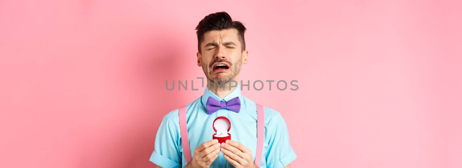 Valentines day. Heartbroken guy crying and holding engagement ring, sobbing from break-up, standing over pink background by Benzoix