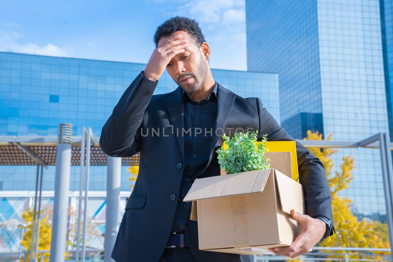 African American businessman fired with cardboard box with office supplies standing sad outside the building. High quality photo