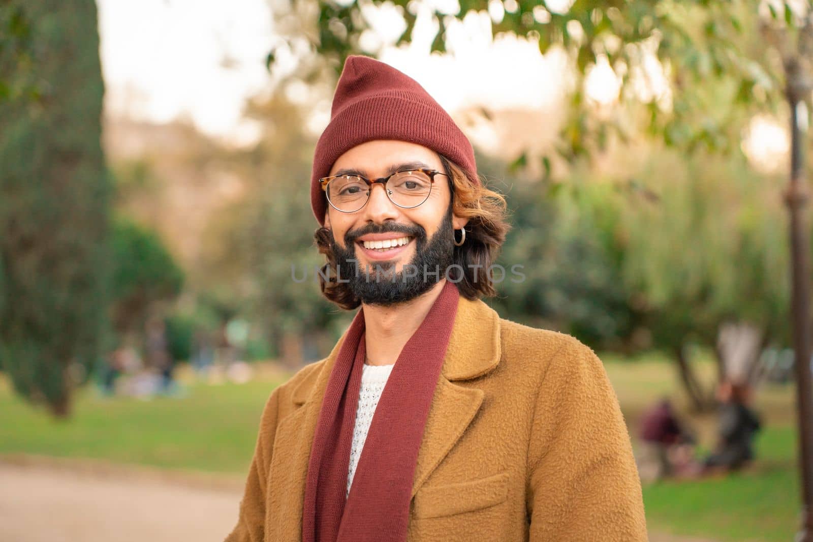 Portrait of a young adult man smiling looking at camera. Hipster man with glasses, beard, earrings, scarf and hat.