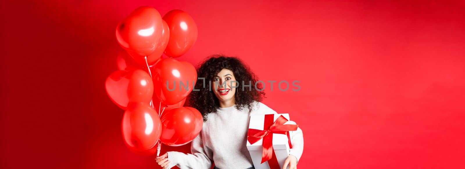 Excited beautiful woman celebrating Valentines day holiday, holding gift box and romantic heart balloons, looking surprised at camera, standing over red background.