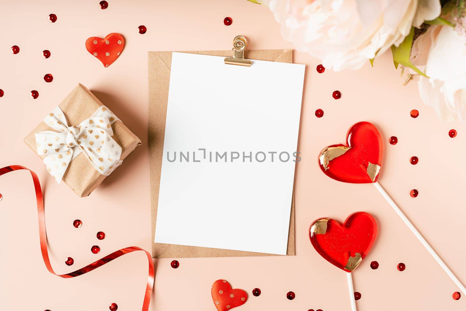 Flat lay of paper envelope with blank mockup greeting paper card. Pink table background with Valentine day gift, letter, heart shape, confetti, lollipops and decoration. Top view, mock up invitations