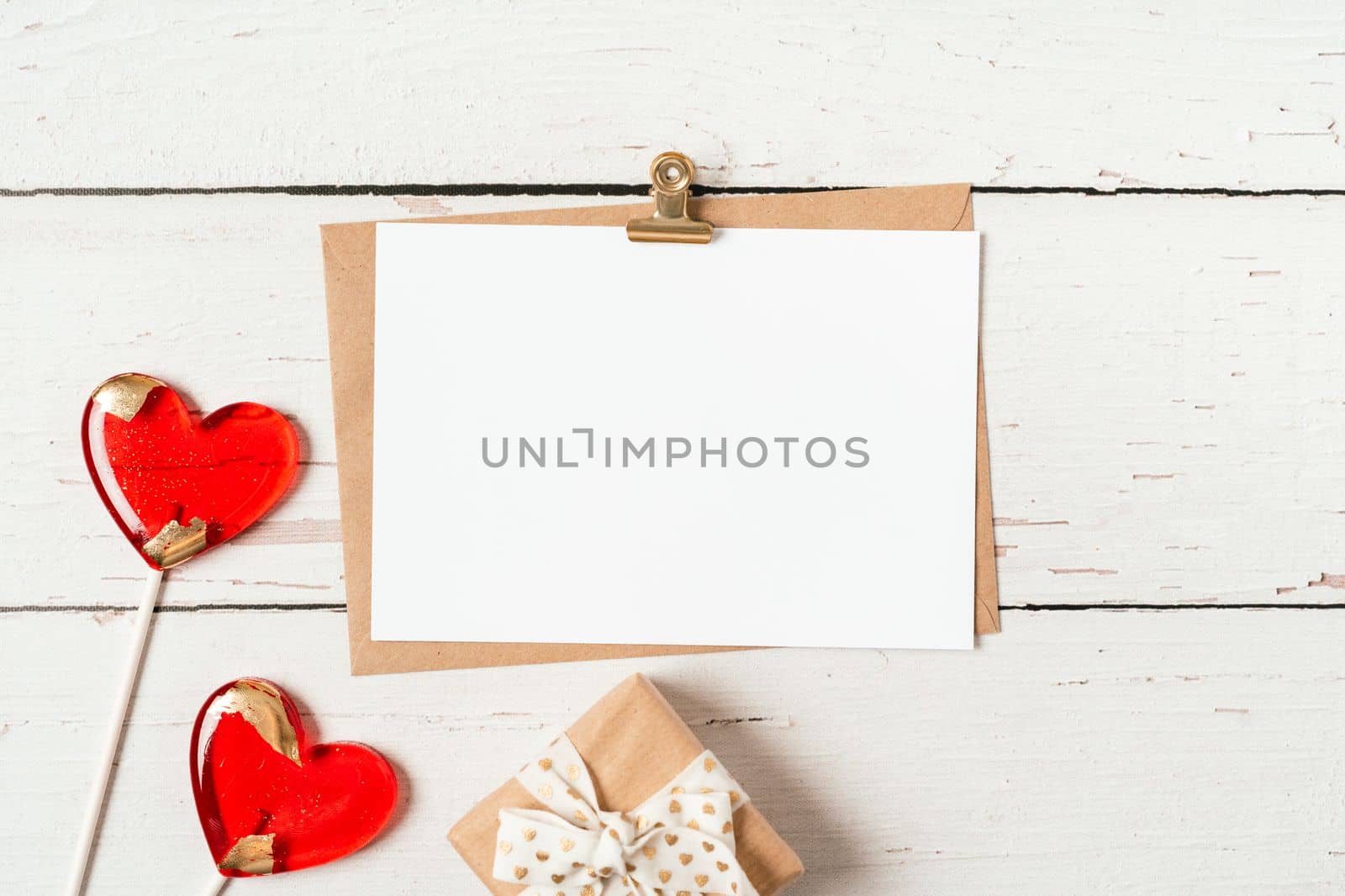 Flat lay of paper envelope with blank mockup greeting paper card. White wooden table background with Valentine day gift, letter, heart shape, lollipops. Top view, mock up invitations