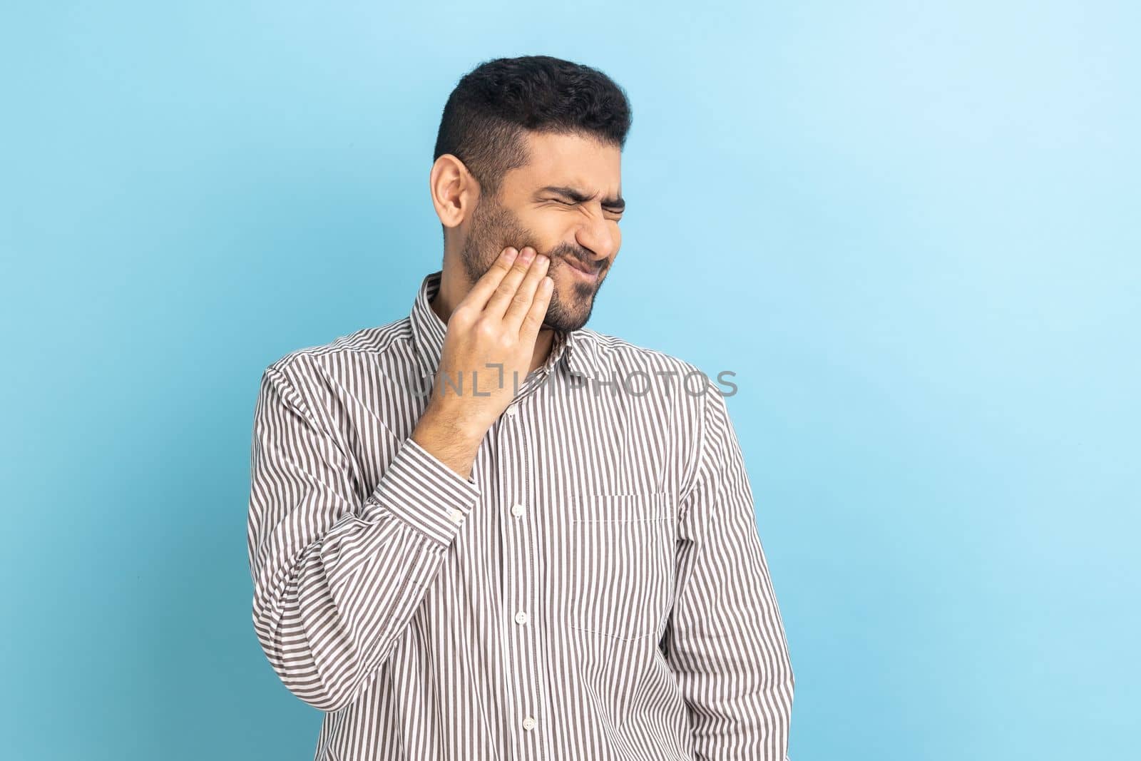 Unhappy bearded businessman feeling toothache, touching sore cheek, suffering from cavities, cracked teeth, gum recession, wearing striped shirt. Indoor studio shot isolated on blue background.