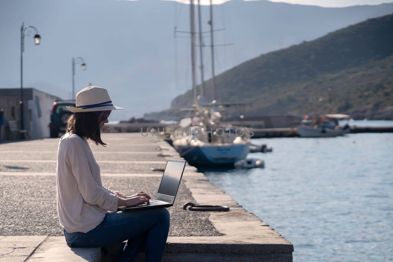 A young girl in a hat is sitting on the pier and working, typing on a laptop keyboard on a sunny day against a beautiful background of a seascape with moored yachts in the bay. Woman works and travels