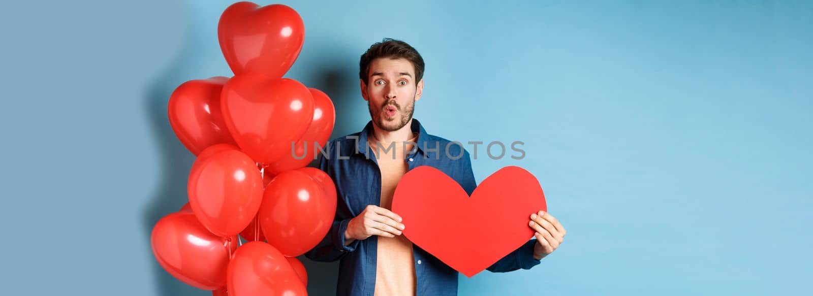 Valentines day concept. Boyfriend in love looking amazed at camera, holding red paper heart and standing with romantic balloon, blue background.