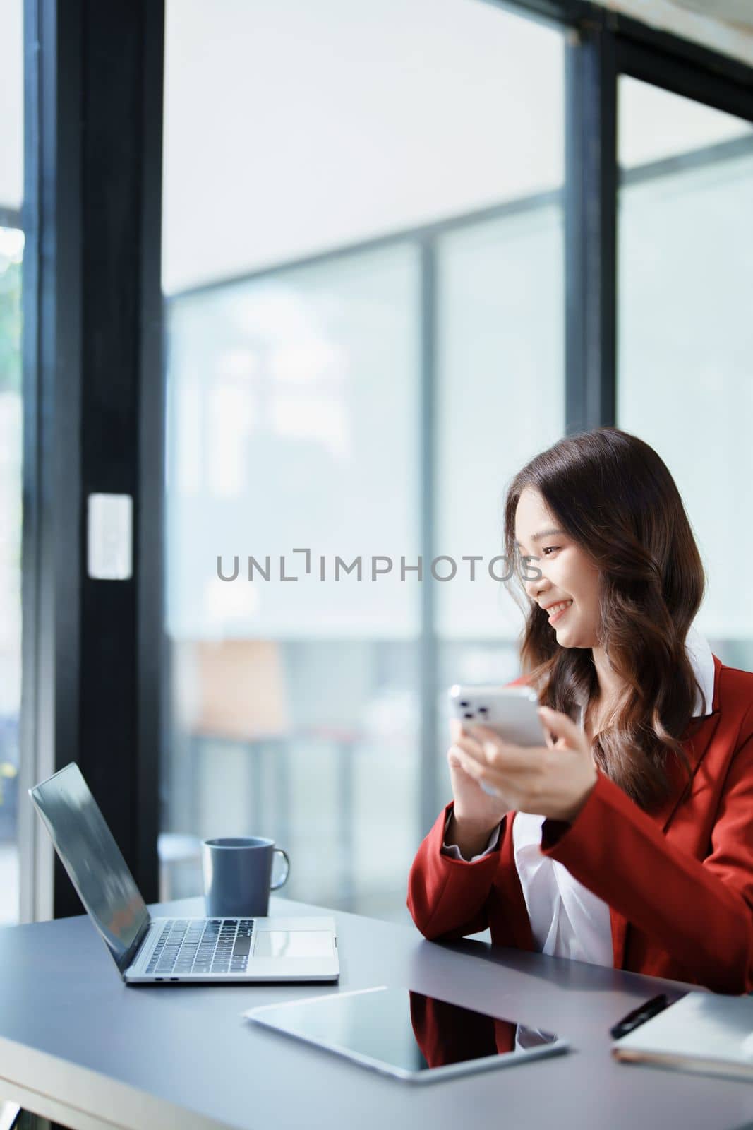 Portrait of a young Asian woman showing a smiling face as she uses her phone, computer and financial documents on her desk in the early morning hours.