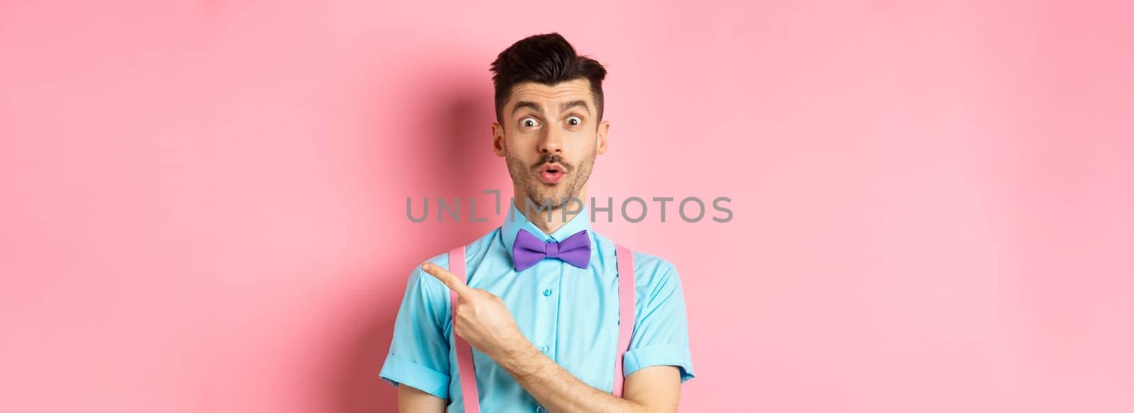 Intrigued and surprised cute guy with moustache showing awesome promo, looking curious and saying wow, pointing finger right at logo, standing on pink background.