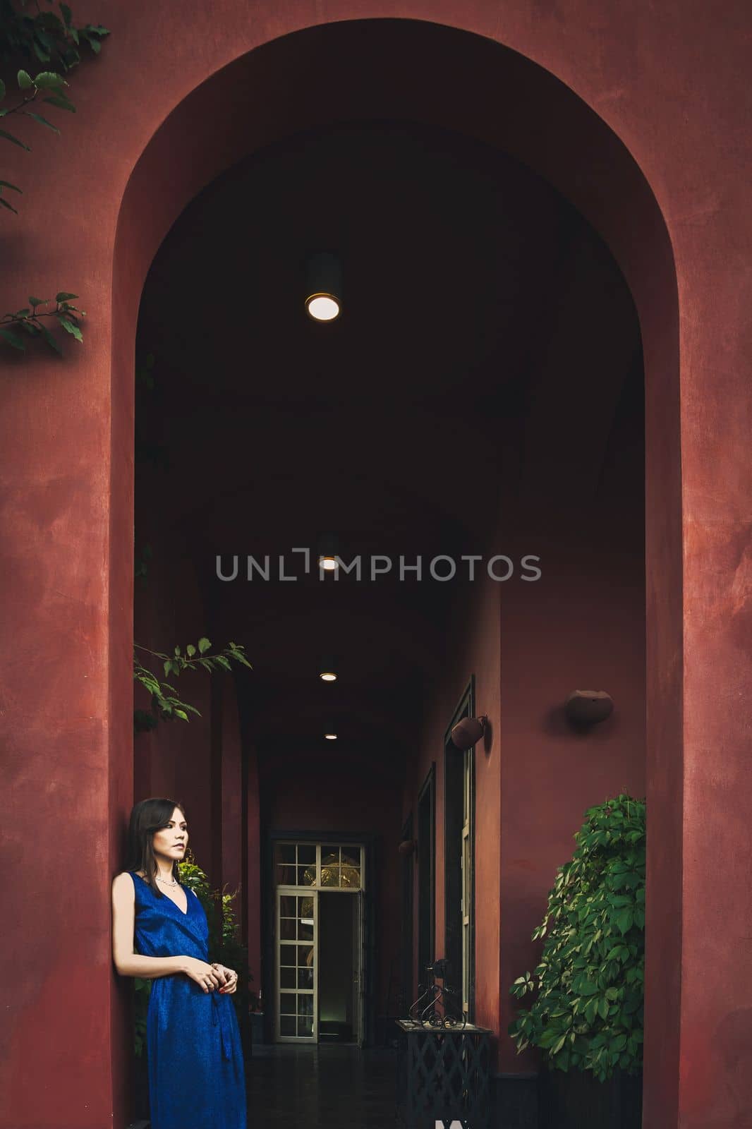 A young beautiful woman in a blue dress stands on a summer terrace under big arch near a large terracotta-colored luxury house.