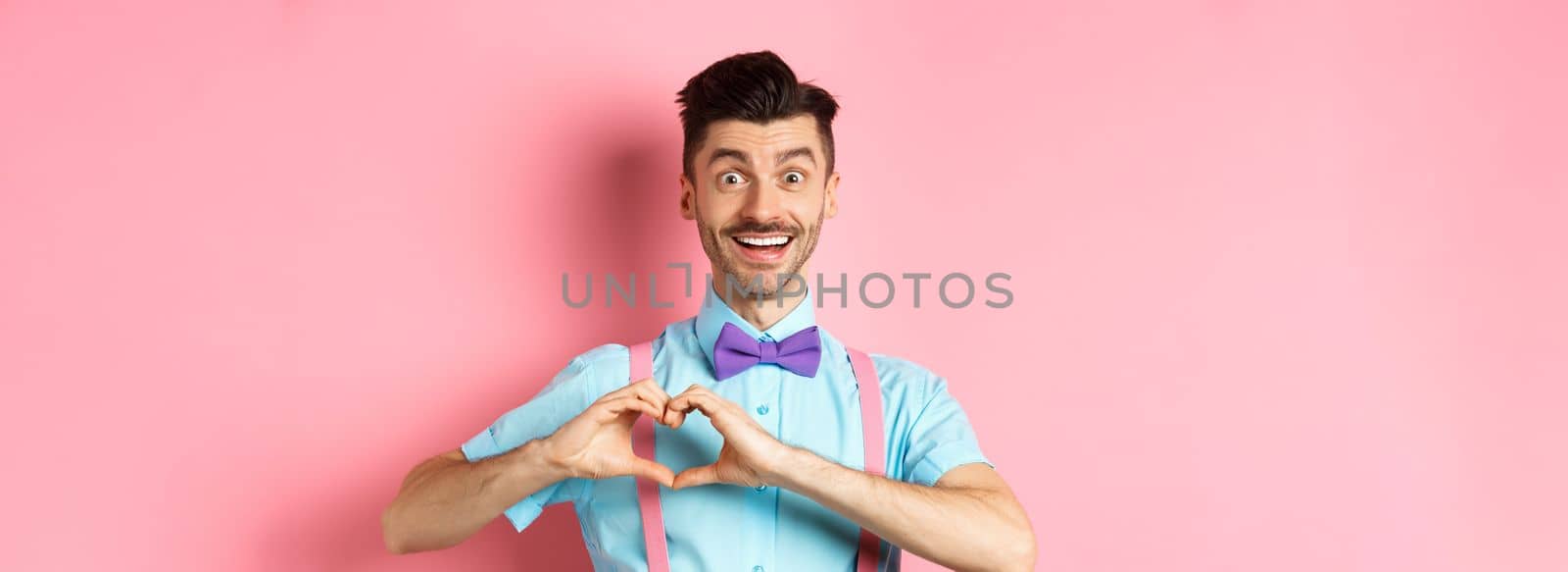 Passionate guy in funny bow tie saying I love you, showing heart gesture on Valentines day and smiling, expressing sympathy to lover, standing over pink background by Benzoix