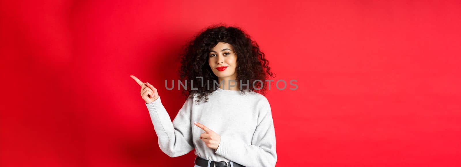 Smiling beautiful girl with curly hair, pointing fingers right at logo, showing advertisement, red background by Benzoix