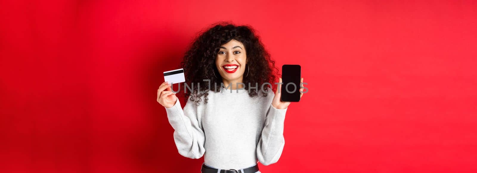 E-commerce and online shopping concept. Cheerful woman smiling, showing plastic credit card and empty smartphone screen, standing on red background.