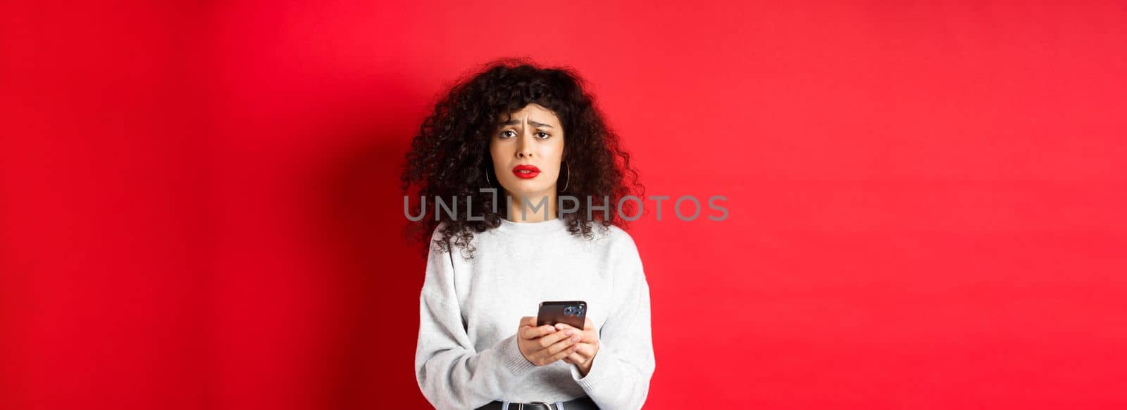 Sad and gloomy woman with curly hair, frowning and feel upset after reading smartphone message, standing disappointed against red background.