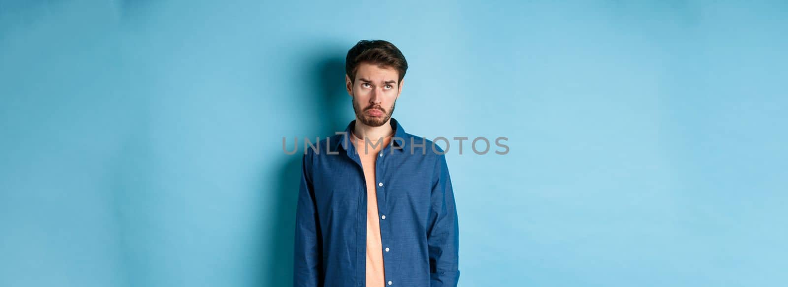 Sad man whining and sulking, looking up with upset face, standing on blue background.