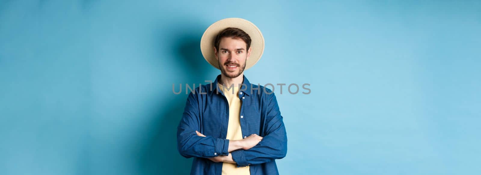 Portrait of cheerful guy going on vacation in straw summer hat, cross arms on chest and smiling, looking confident, standing on blue background.