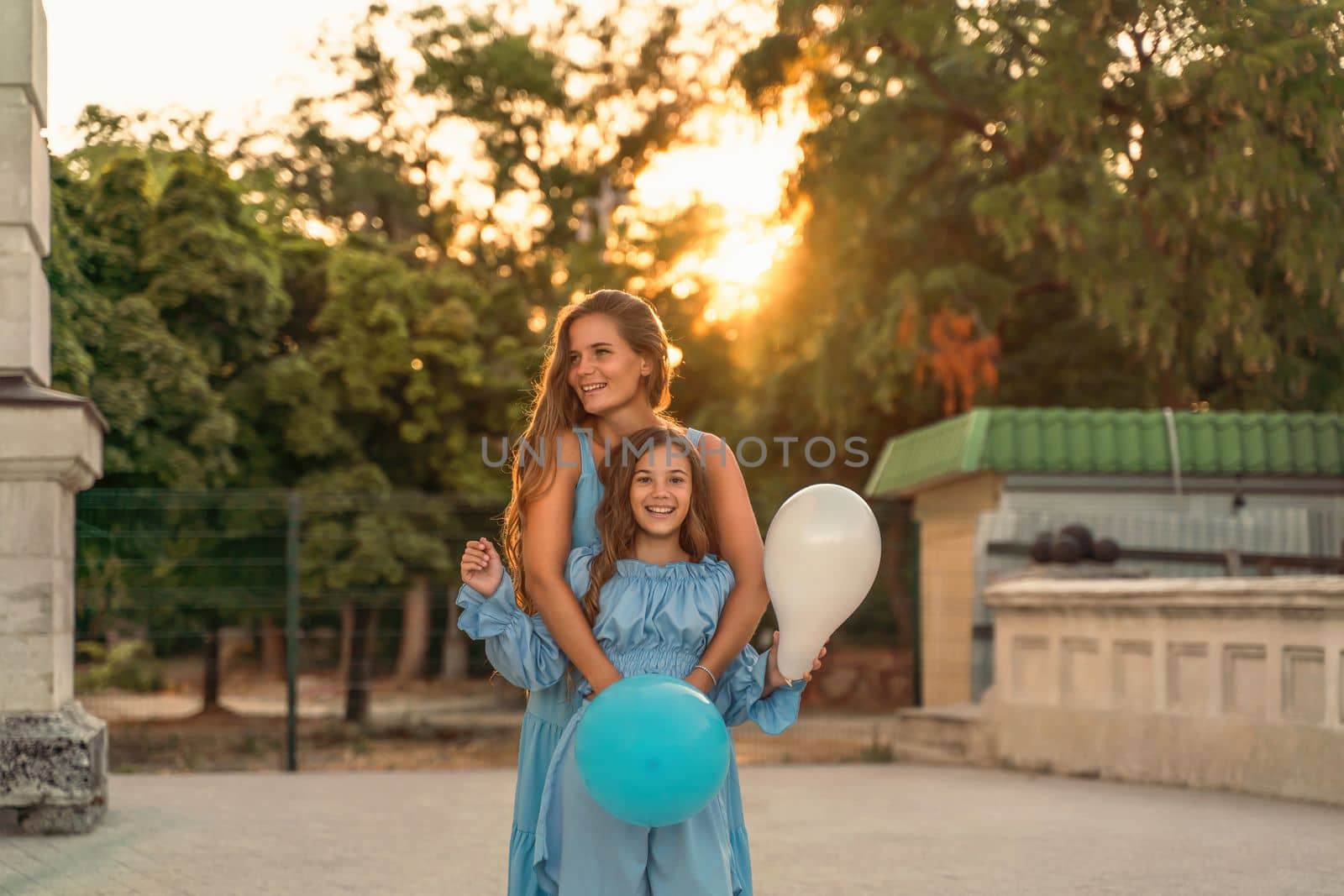 Mother daughter family sunset. Portrait of mother and daughter in blue dresses with flowing long hair against the backdrop of sunset. A woman hugs and presses the girl to her, holding balloons in her hands by Matiunina