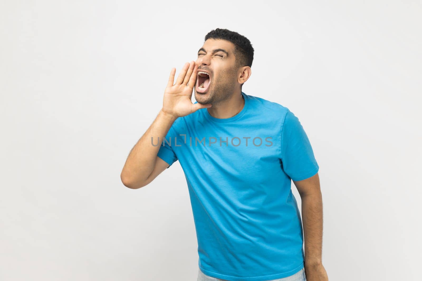 Portrait of excited handsome attractive unshaven man wearing blue T- shirt standing keeps hands near opened mouth, screaming news. Indoor studio shot isolated on gray background.