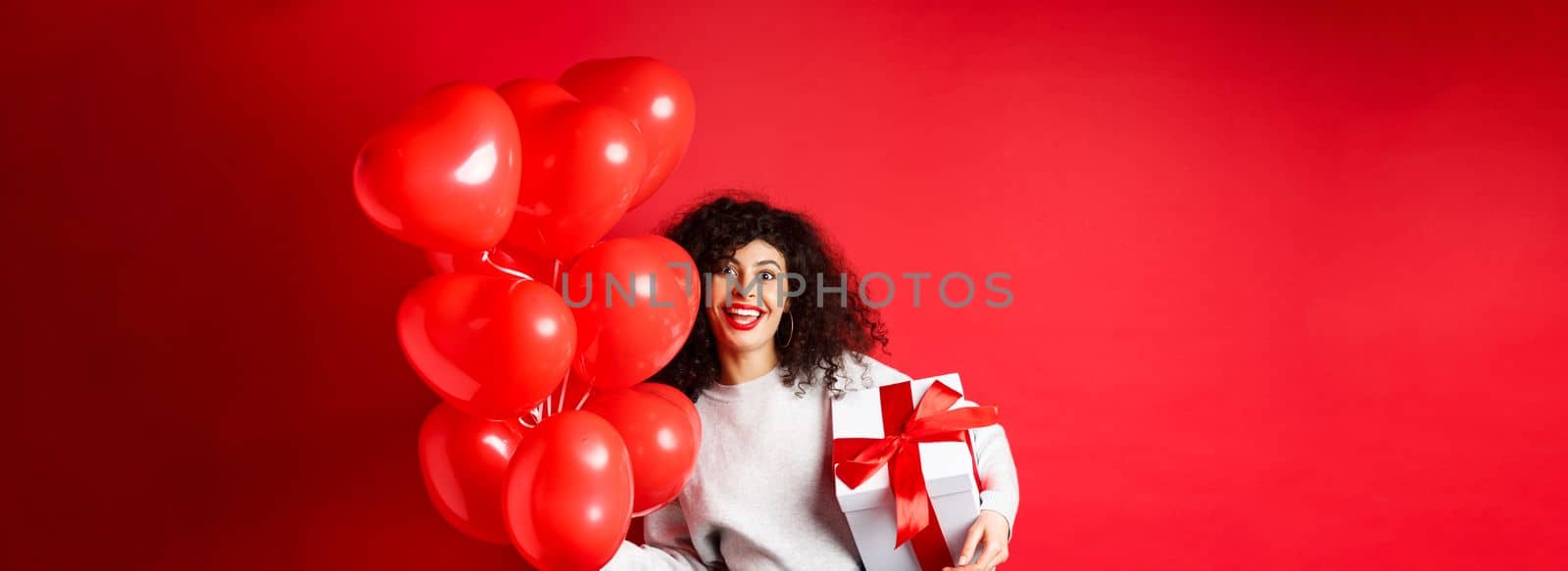 Holidays and celebration. Happy birthday girl holding gift and posing near party helium balloons, smiling excited at camera, red background.