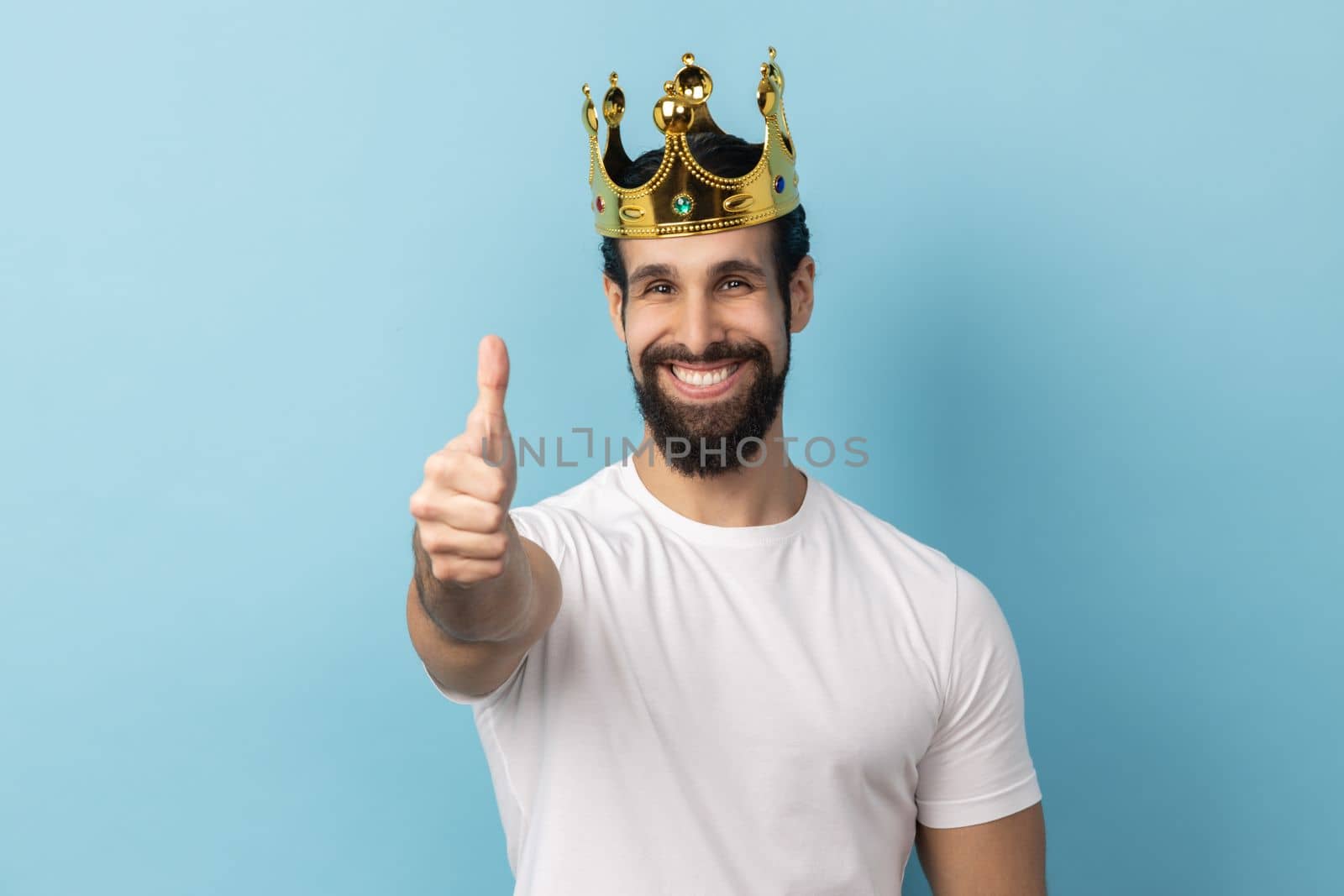 Portrait of smiling joyful man with beard wearing white T-shirt and in gold crown standing looking at camera with toothy smile, showing thumb up. Indoor studio shot isolated on blue background.
