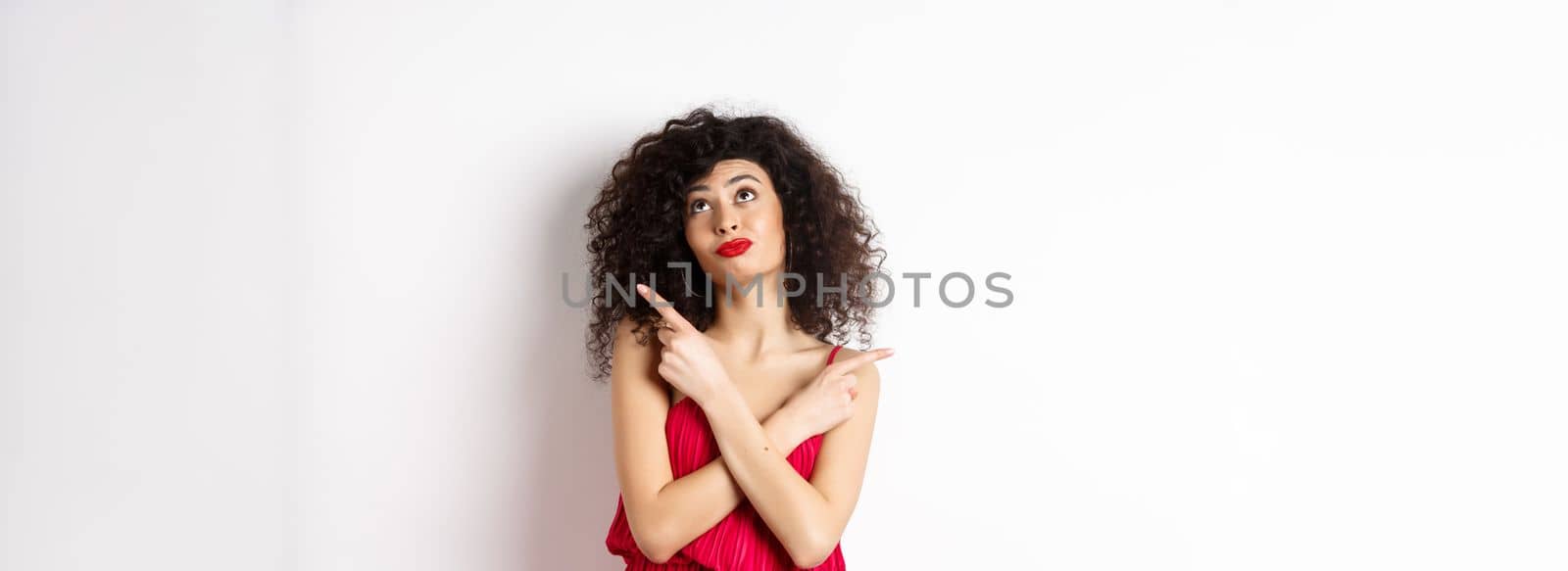 Indecisive silly woman in red dress, cross fingers and pointing sideways, looking up pensive, making choice, standing over white background.