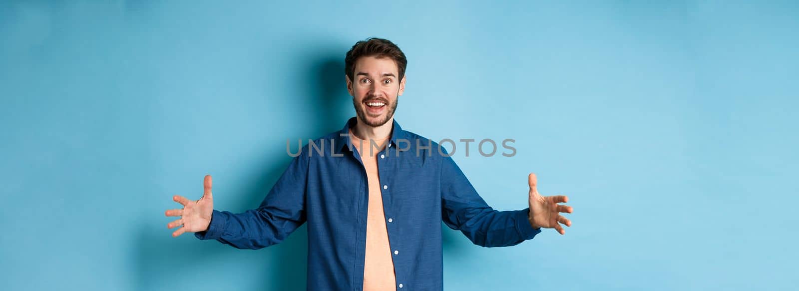 Happy smiling man spread hands sideways and showing big size object, talking about large object, standing cheerful on blue background.