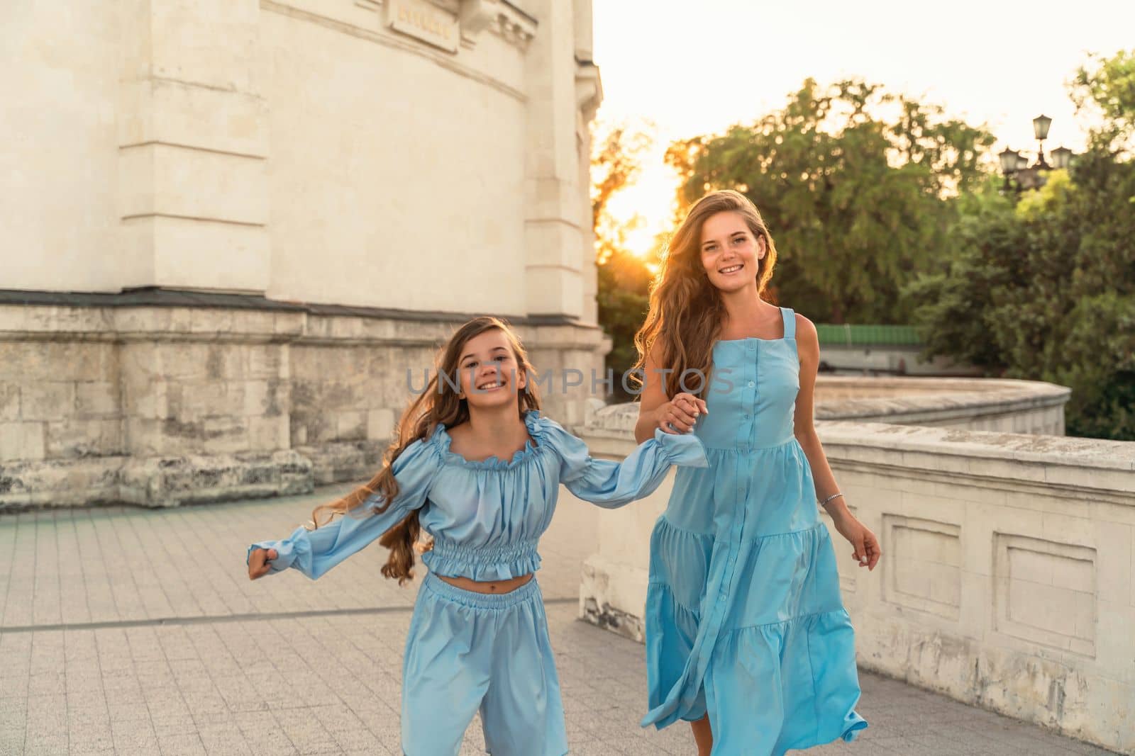 Portrait of a mother and daughter in blue dresses with flowing long hair against the backdrop of a sunset and a white building. They run and smile at the camera. Family stories on the weekend.