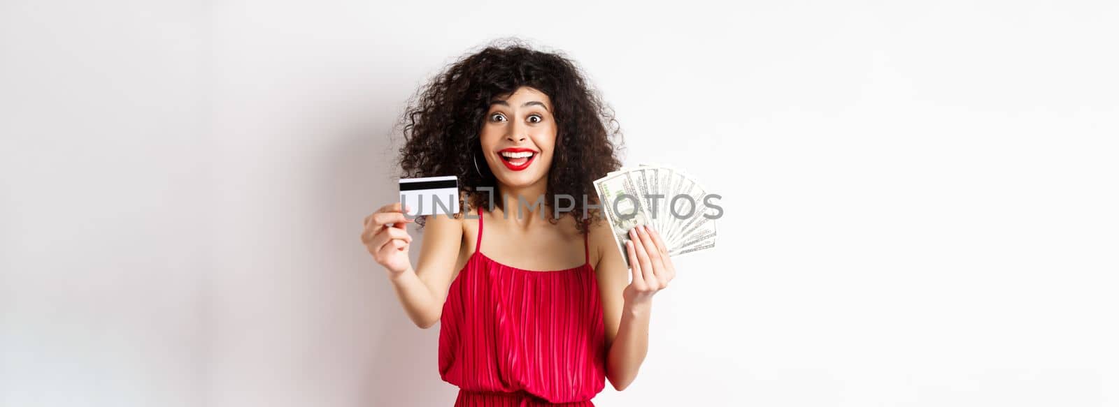 Shopping. Excited curly-haired woman in red dress, holding money but showing plastic credit card with happy smile, white background.
