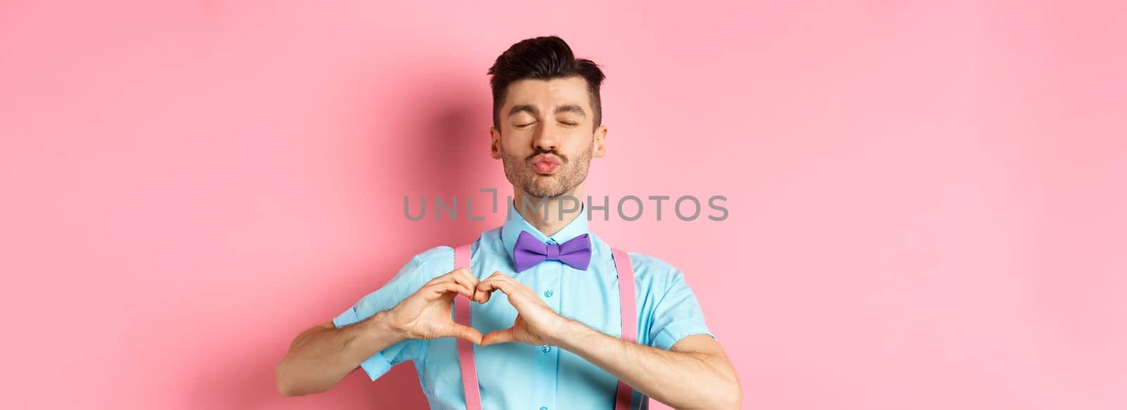 Romantic man in funny bow-tie waiting for kiss, close eyes and pucker lips kissing, showing heart gesture, being in love on Valentines day, standing over pink background.