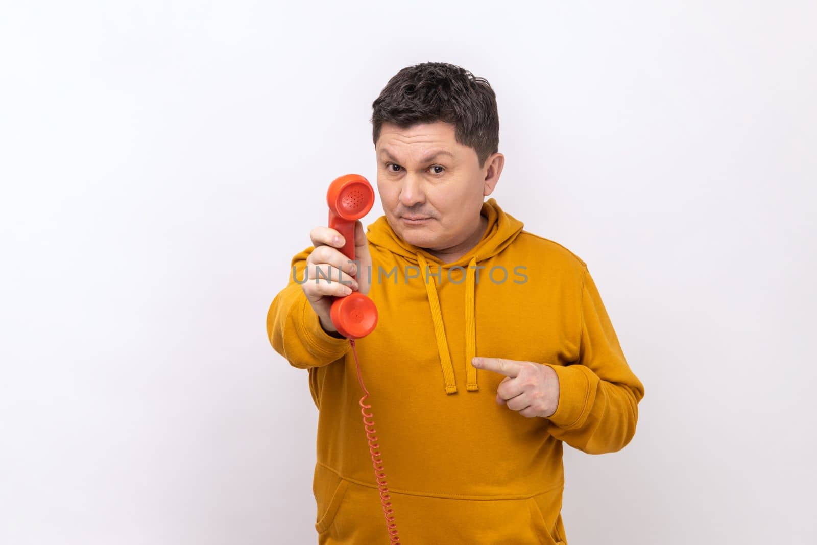 Middle aged man pointing finger at handset of red vintage landline phone, interested in retro devices, answering calls, wearing urban style hoodie. Indoor studio shot isolated on white background.
