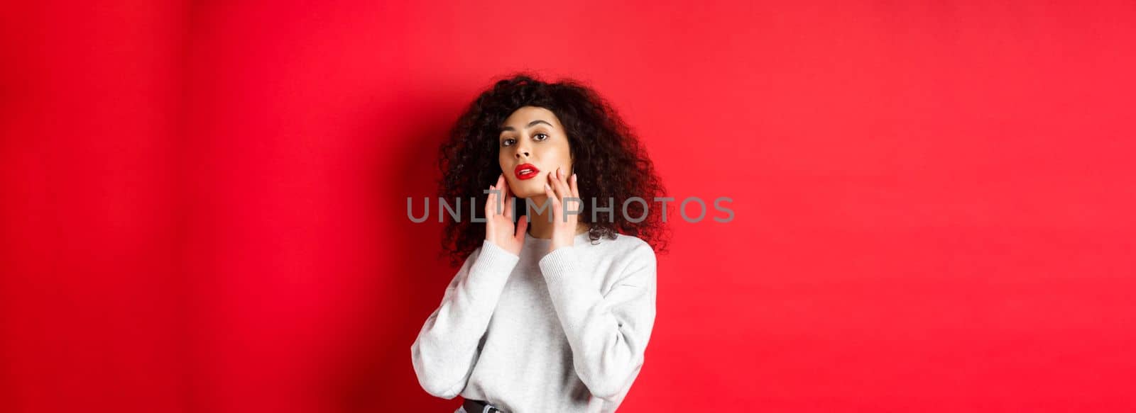 Beautiful and stylish curly woman with red lips, touching perfect young face and looking sensual at camera, standing against red background by Benzoix